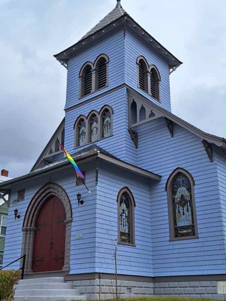 Exterior view of a lavender-colored church with stained-glass windows, featuring a rainbow Pride flag on the front.