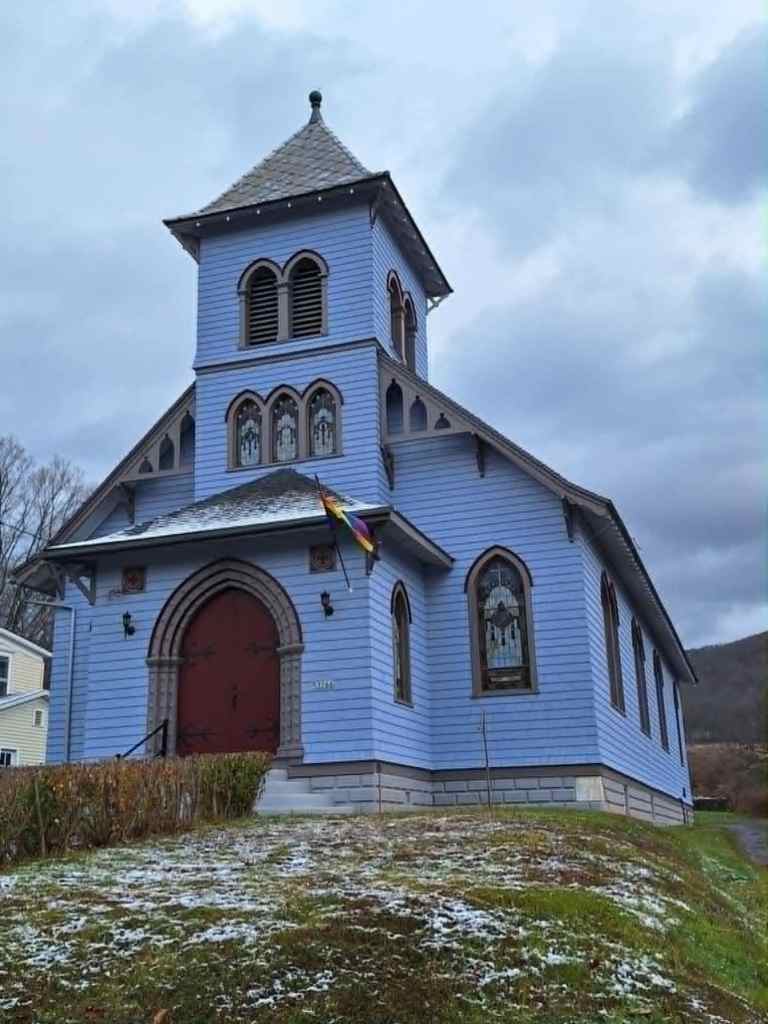 A lavender-painted former church with a cross on its facade, featuring stained glass windows, and a rainbow Pride flag flying outside.