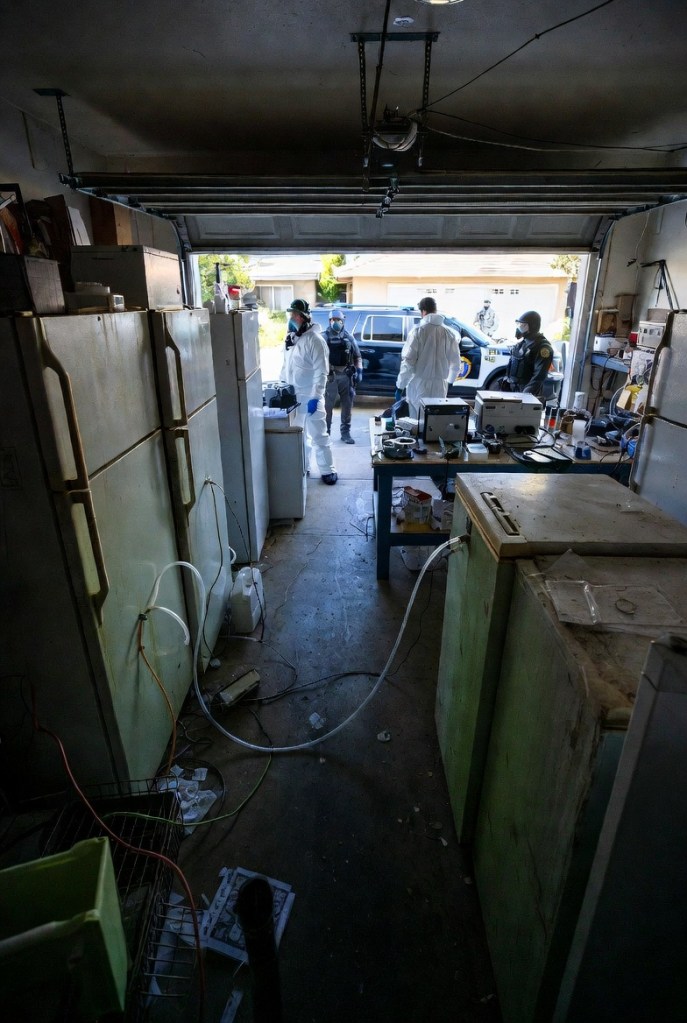 A dimly lit garage with a view of the driveway, featuring several refrigerators and various equipment. People in protective clothing are discussing, while law enforcement officers observe from outside.