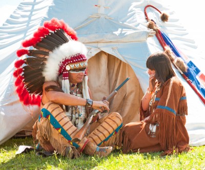 A Native American man wearing a feathered headdress and traditional attire sits with a rifle, while a woman in a dress with fringes sits next to him, in front of a tipi.