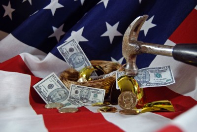 A hammer striking a nest filled with money, including American dollar bills and gold coins, placed on a backdrop of the American flag.