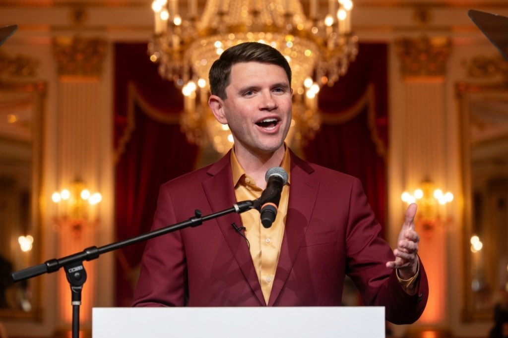 A man in a maroon blazer and gold shirt speaks enthusiastically at a podium, with a microphone, in an elegantly decorated room featuring chandeliers and rich drapery.