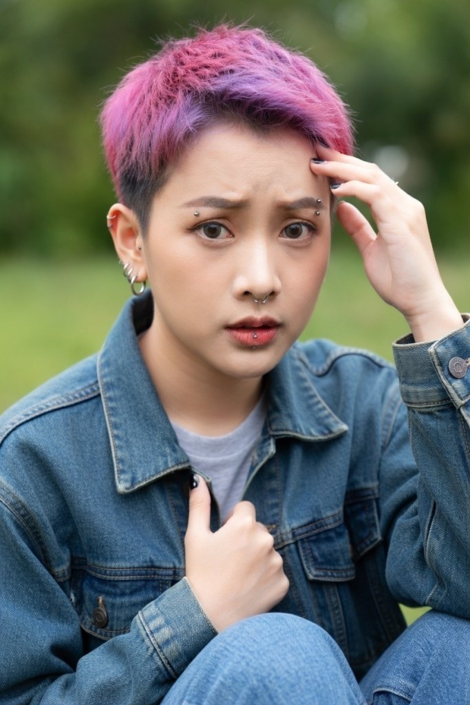 A young person with short, dyed pink and purple hair sitting outdoors, looking concerned while holding their head with one hand.