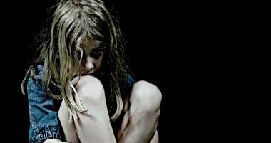 A young girl sitting on the floor, hugging her knees with a distressed expression, set against a dark background.