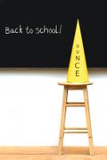 A yellow dunce cap placed on a wooden stool in front of a black chalkboard that says 'Back to school!'