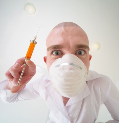 A person wearing a white shirt and a face mask, holding a syringe with orange liquid, looking directly at the camera with an intense expression.