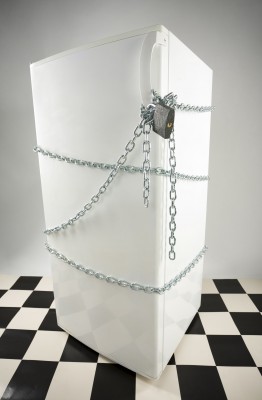 A white refrigerator wrapped in chains and locked with a padlock, standing on a black and white checkered floor.