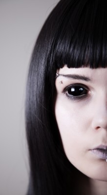 Close-up of a woman with dark hair and striking makeup, featuring black eye contacts and a lip piercing.