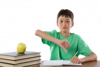 A young boy wearing a green shirt gives a thumbs down gesture while sitting at a desk with an open notebook and a green apple on a stack of books.