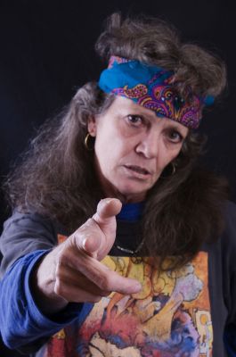 A woman with long hair wearing a colorful headband and a graphic t-shirt, pointing towards the camera with an intense expression.